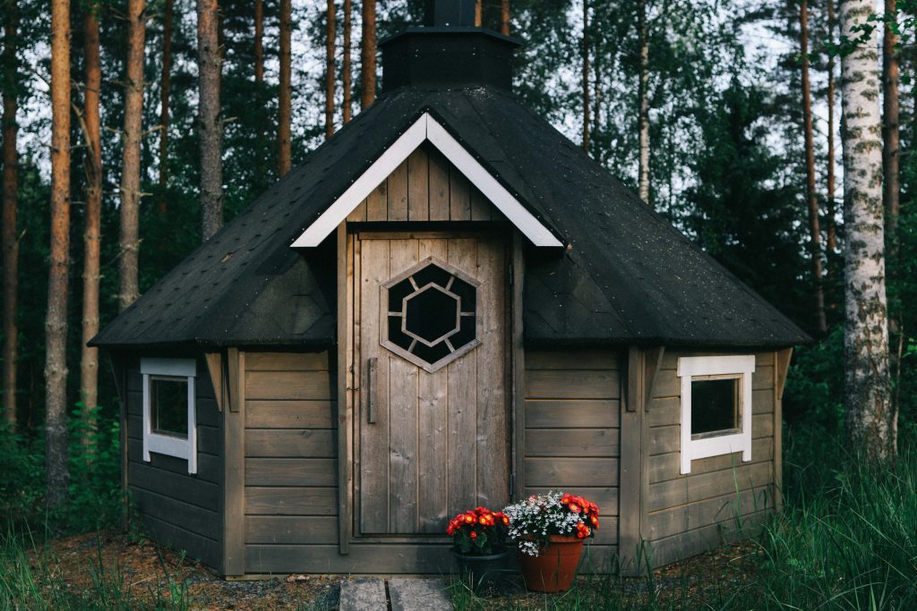 A small log cabin in the middle of the woods in Tampere. Has a small chimney and white windows.